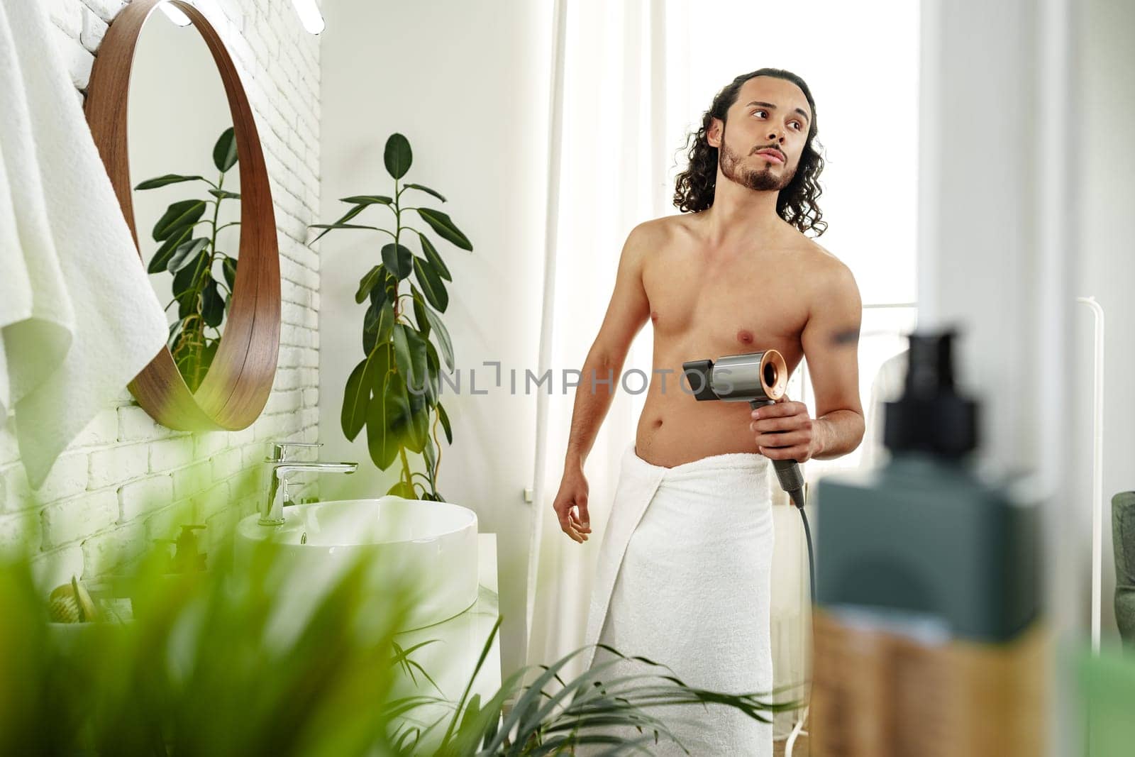 A man with curly hair styles himself in a bright room while using a hairdryer to enhance his look close up