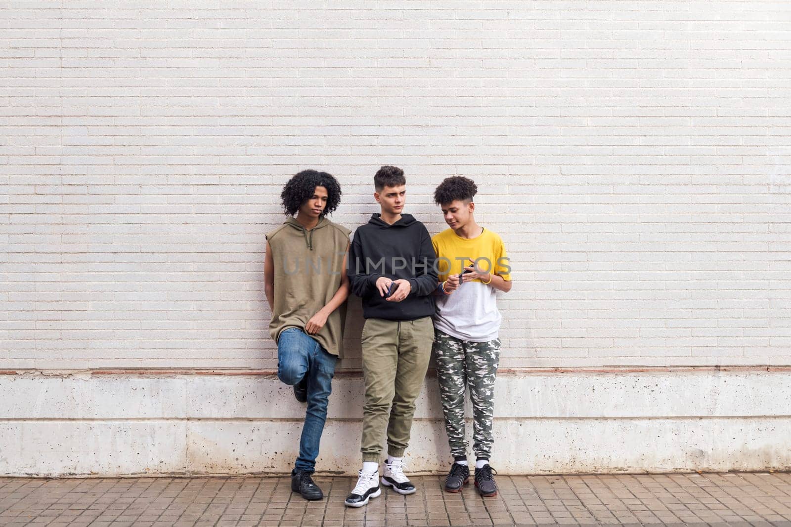 full body of three multiethnic teenager friends leaning against white brick wall, concept of friendship in youth and urban lifestyle, copyspace for text