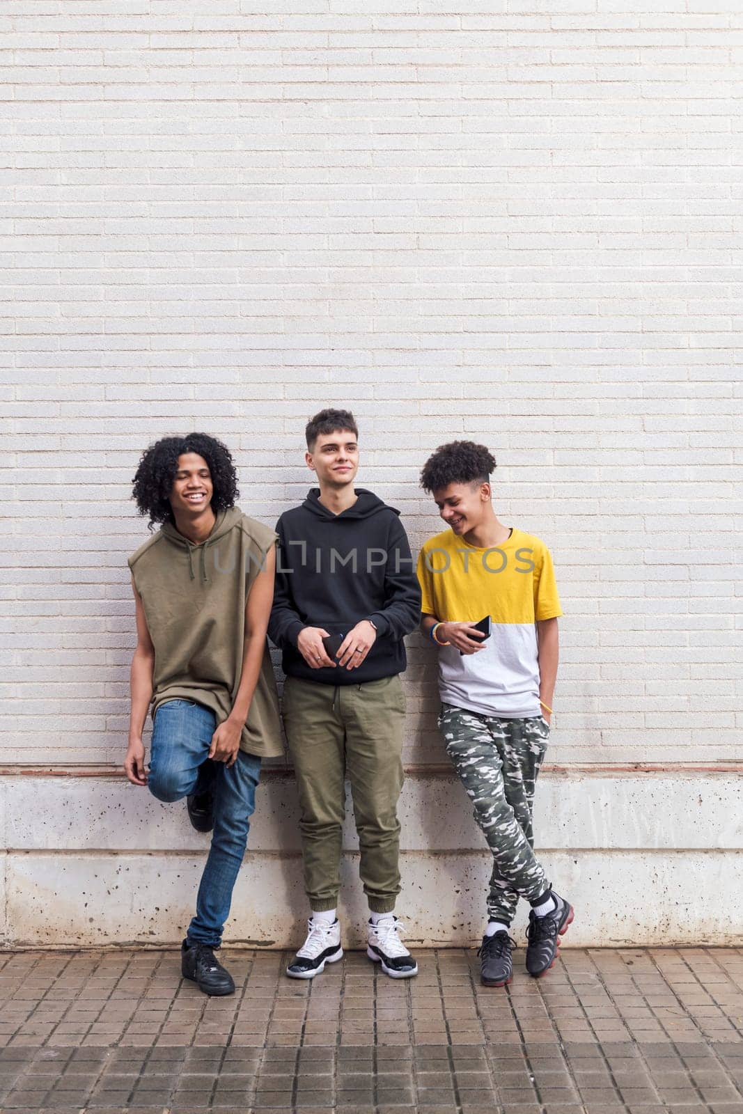 full body of three multiethnic teenager friends leaning against white brick wall, concept of friendship in youth and urban lifestyle, copyspace for text