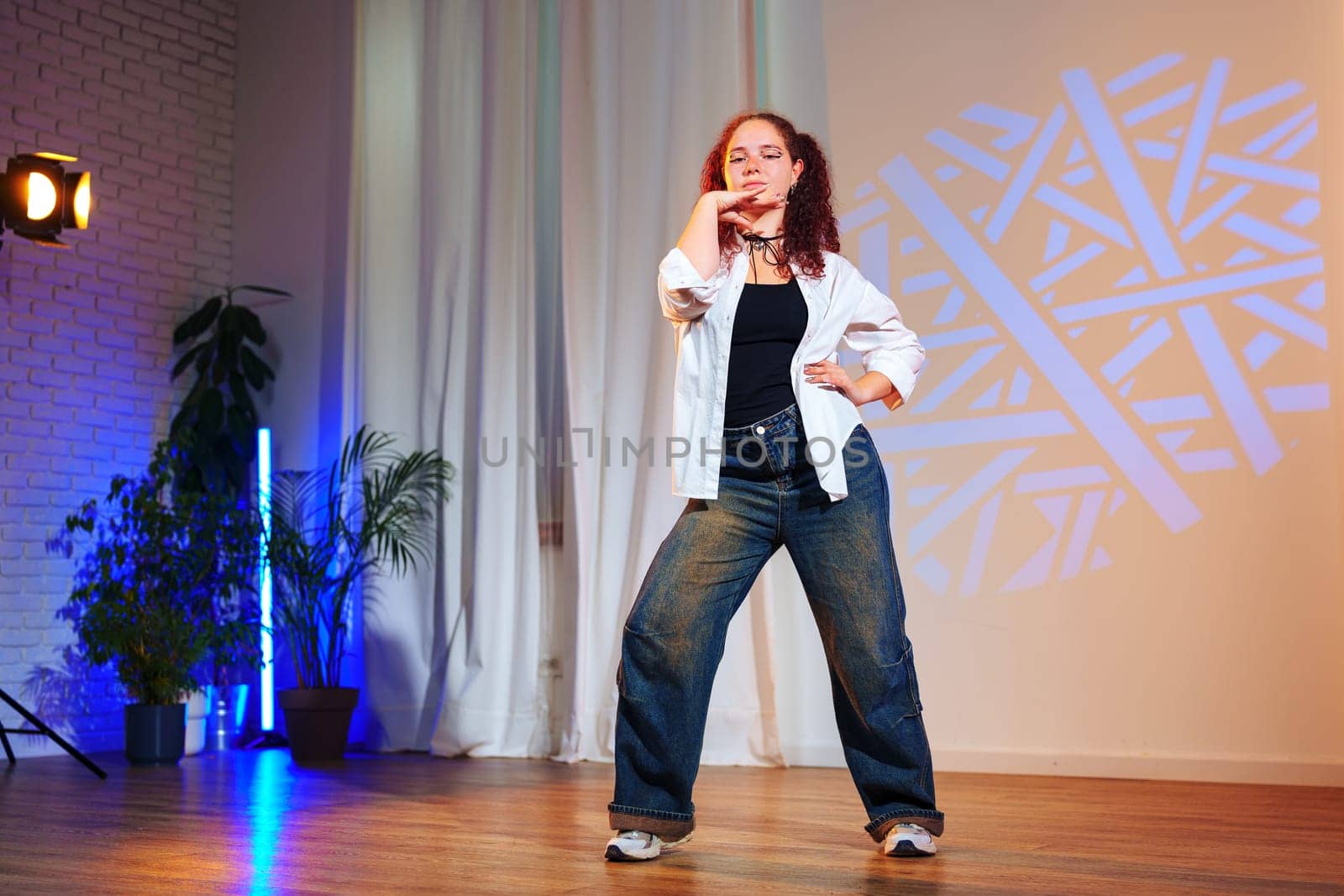 A young woman with curly hair, wearing a white shirt and blue jeans, strikes a pose on a dance floor. She is standing in front of a white curtain and a projection of blue geometric shapes.