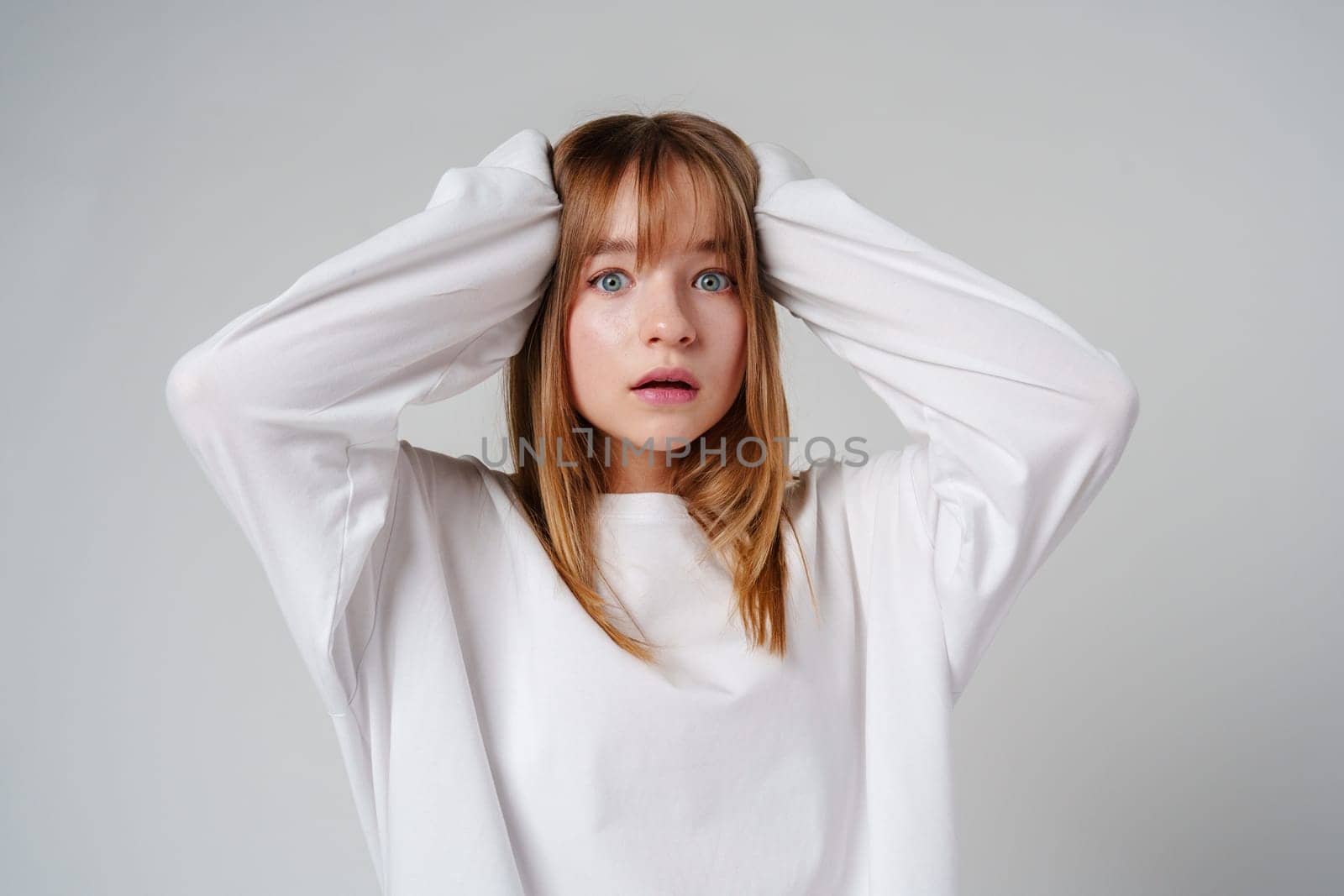 A teen girl in a loose white sweater looks surprised with her hands on her head against a plain gray backdrop close up