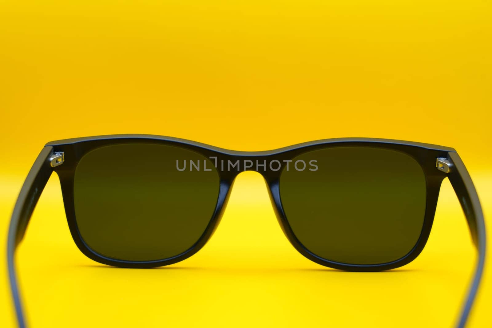 A close-up back view of black sunglasses with dark lenses, displayed against a vibrant yellow background, emphasizing minimalist design and summer style.