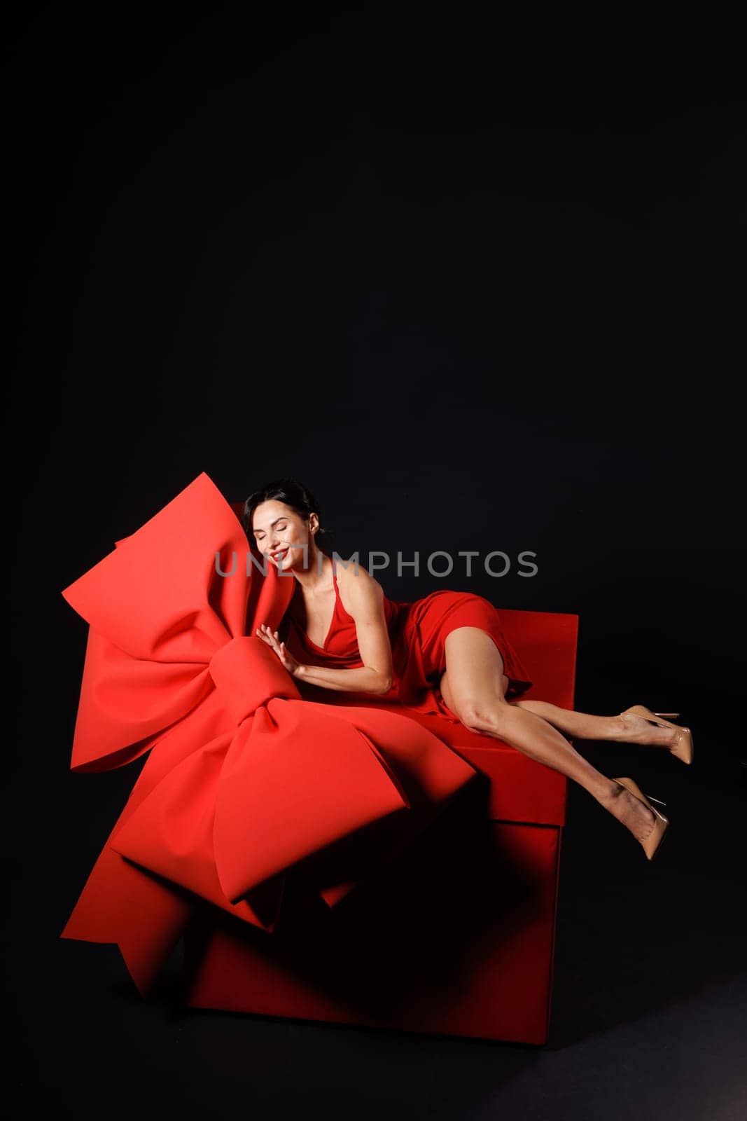 girl with big red bow, sitting on huge red gift box, on dark background, surprise, trend,