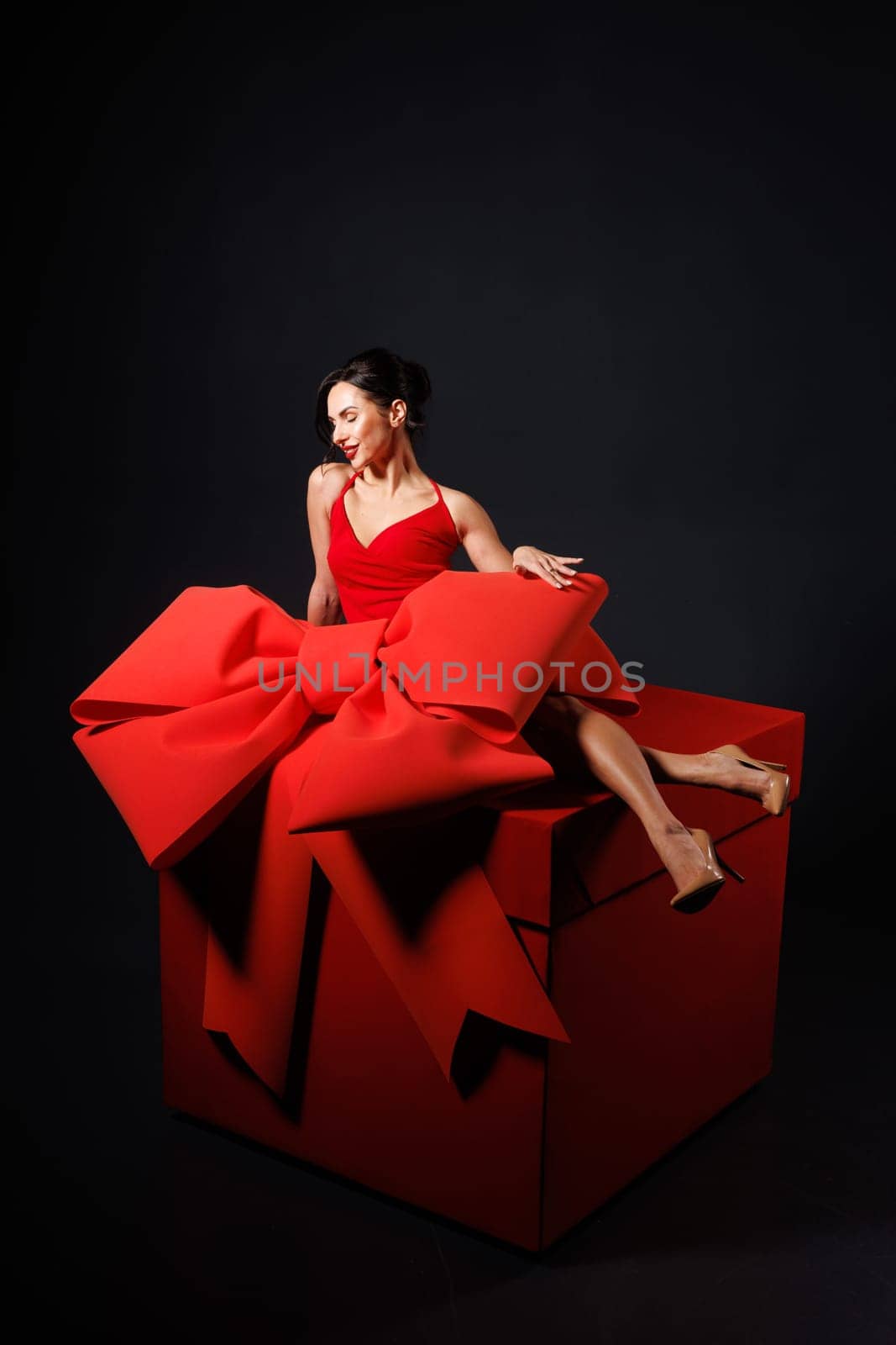 girl with big red bow, sitting on huge red gift box, on dark background, surprise, trend,