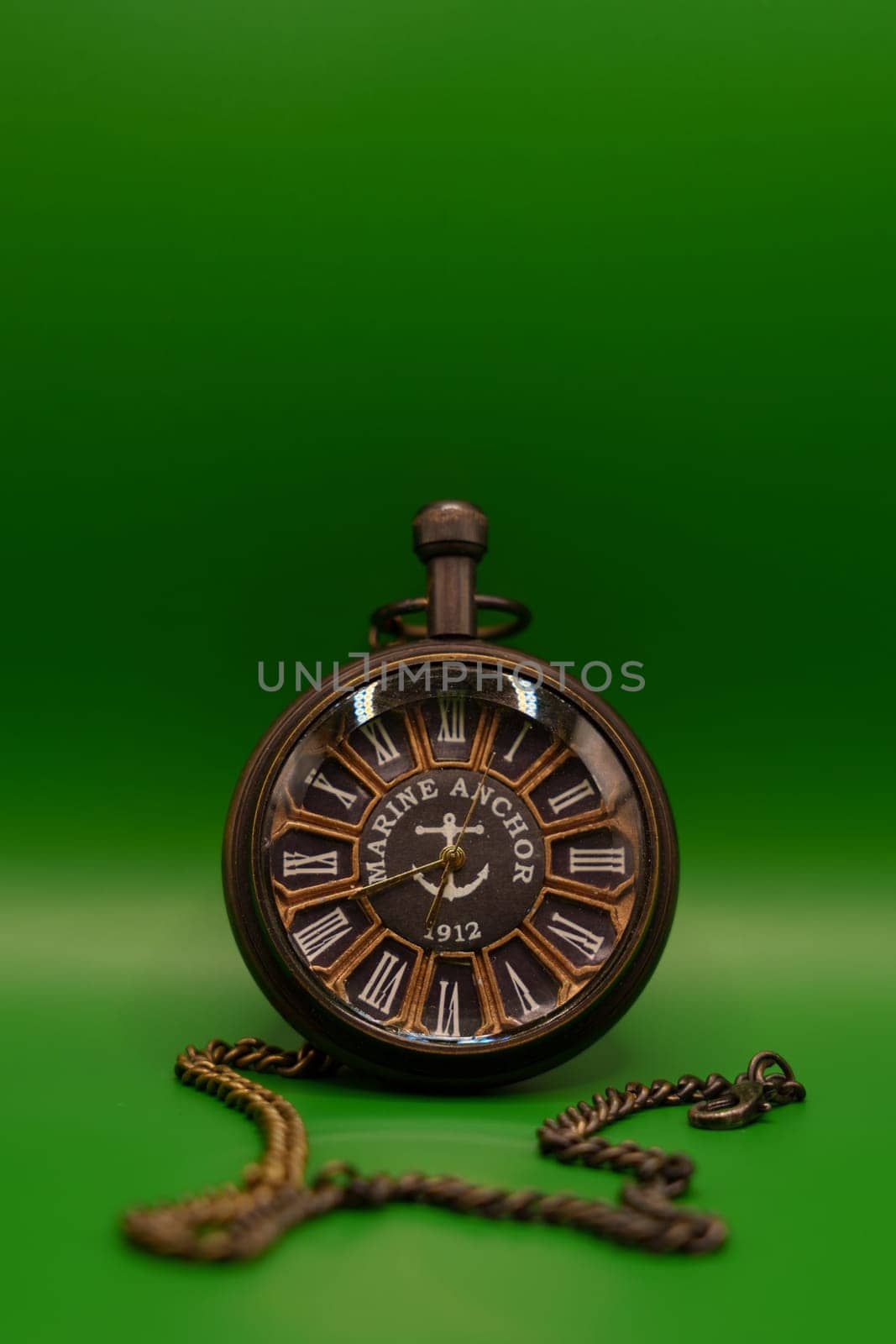 A vintage pocket watch featuring Roman numerals and "Marine Anchor 1912" design, displayed with a chain on a solid green backdrop.