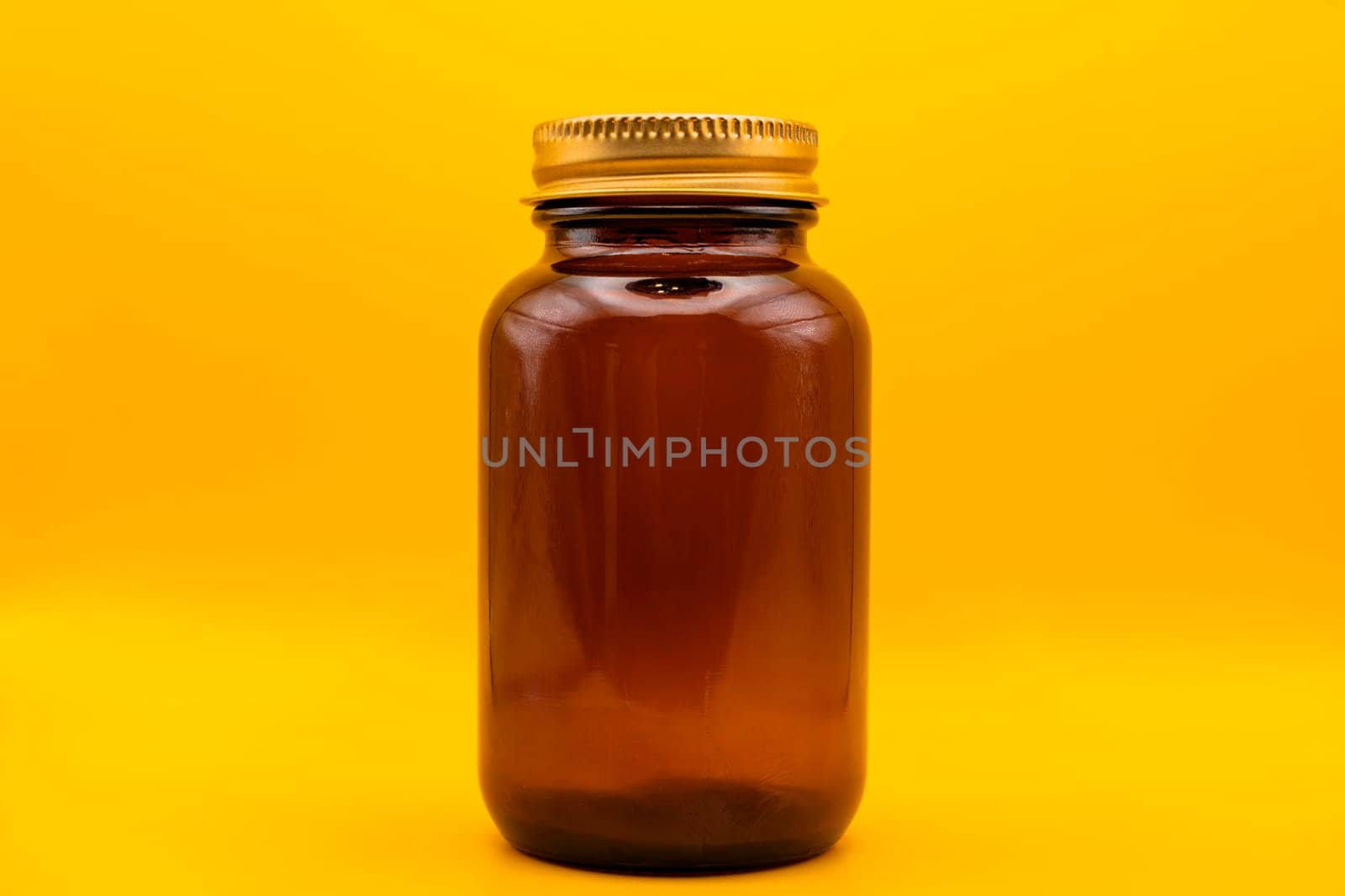 A close-up of an empty amber glass jar with a golden lid, isolated on a vibrant yellow background, perfect for branding or packaging concepts.