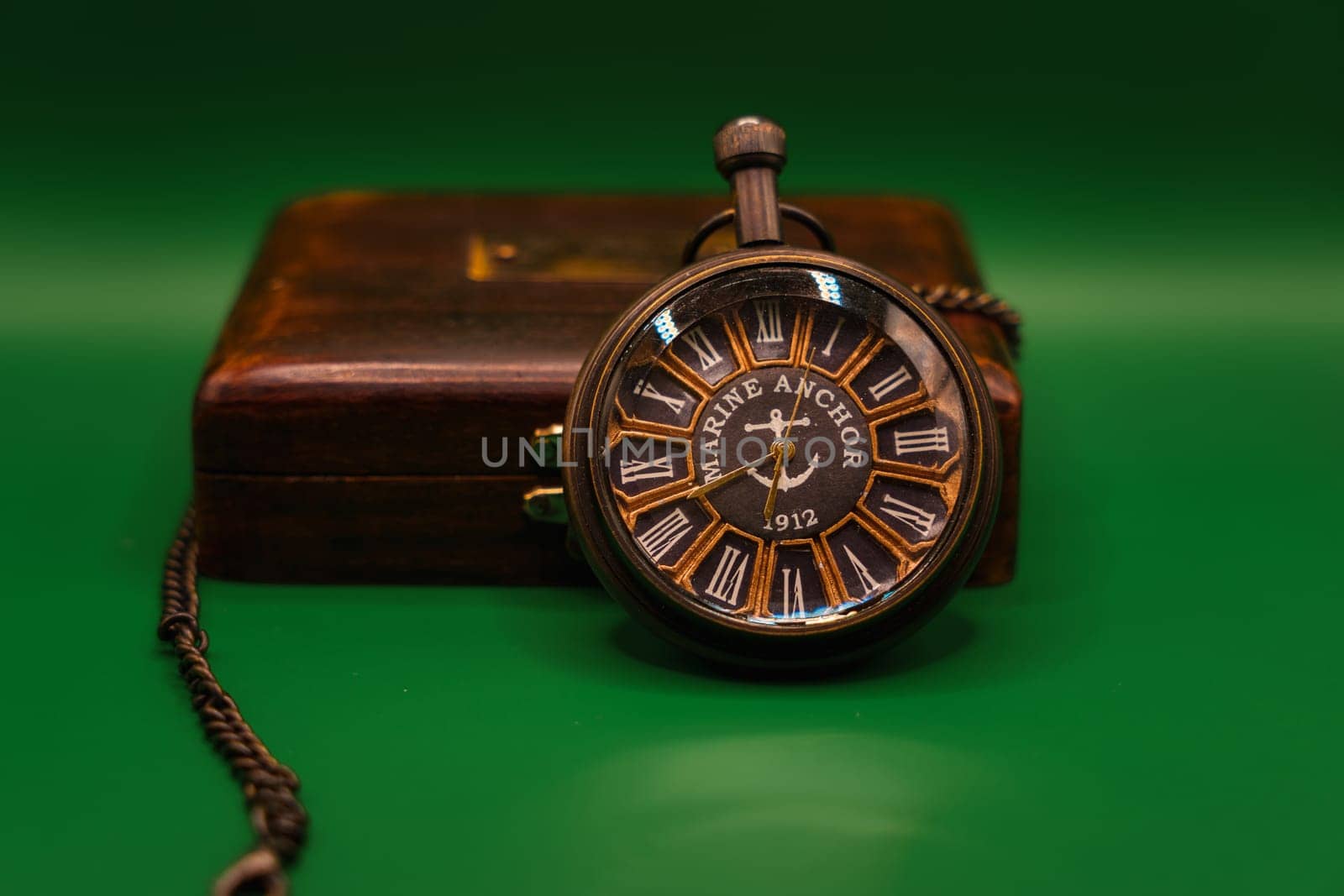 Vintage pocket watch marked "Marine Anchor 1912" with chain displayed on a wooden box, photographed against a green background.