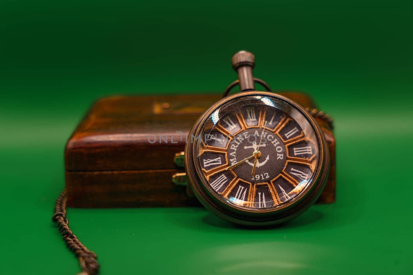 Antique pocket watch labeled "Marine Anchor 1912" with Roman numerals, placed on a vintage wooden box, set against a green background.