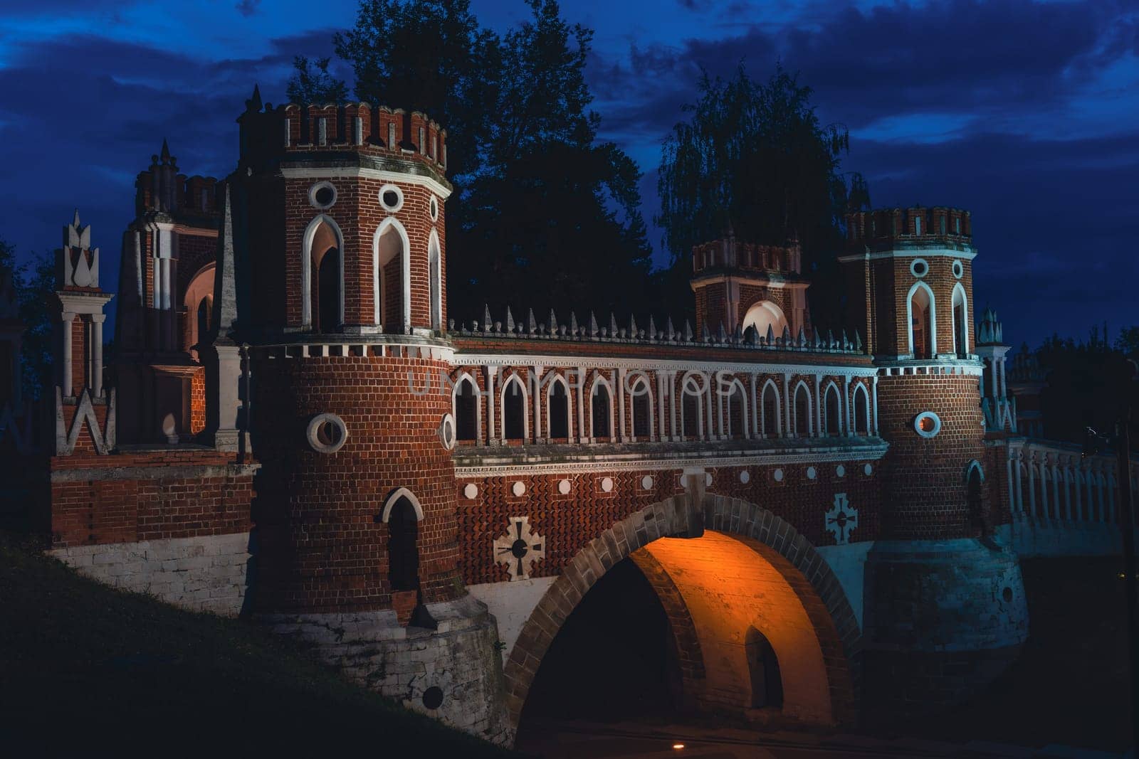 A Gothic-style brick bridge with decorative towers, pointed arches, and illuminated details, captured under a dark night sky.