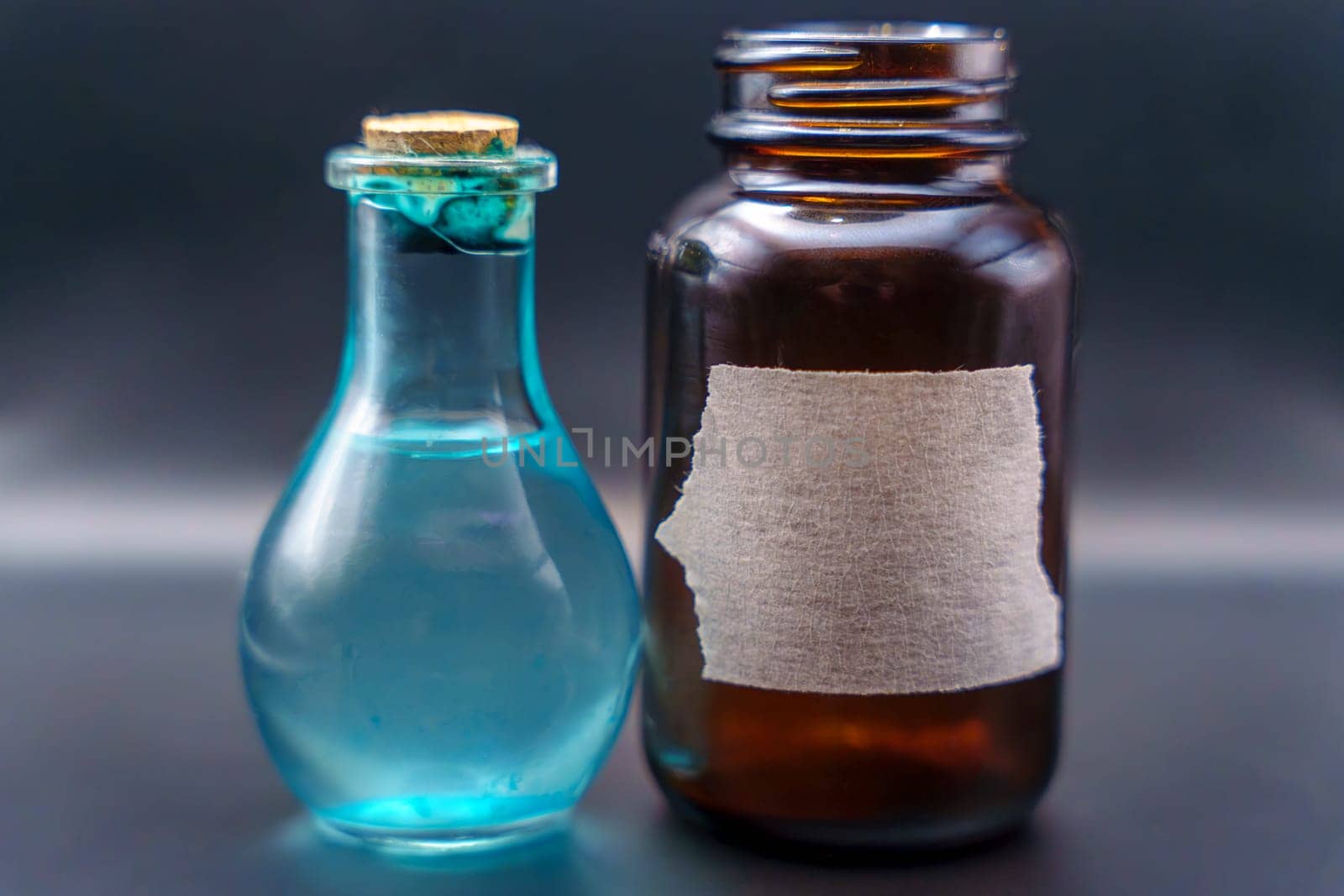Close-up of a brown bottle with a torn label and a corked blue glass bottle containing liquid, set against a dark background.