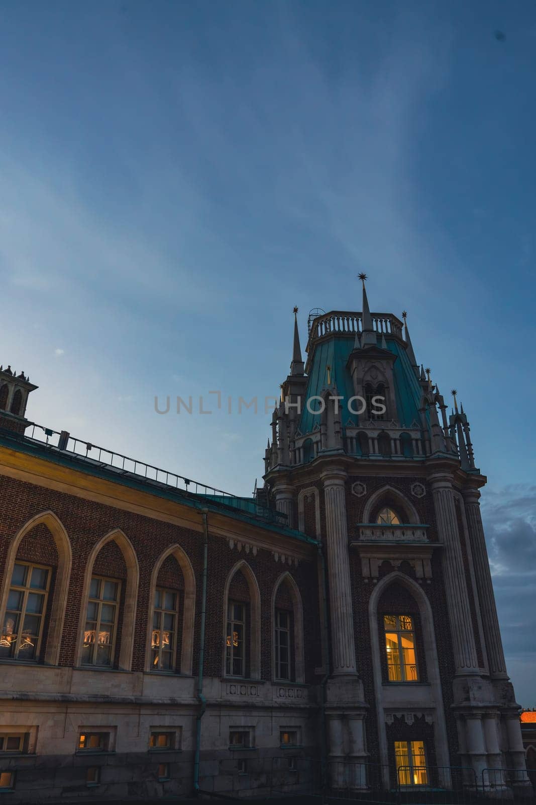 Close-up of a Gothic-style palace tower with spires and illuminated arched windows, captured under a serene twilight sky.