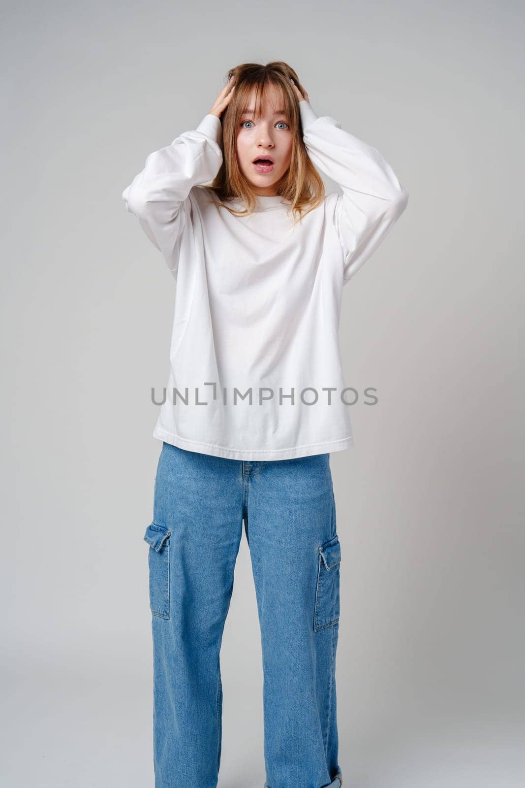A young woman with light brown hair and blue eyes expresses surprise in a minimalist studio setting
