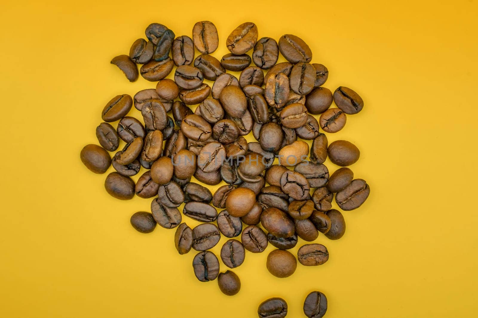 Overhead view of a heap of roasted coffee beans spread on a vibrant yellow background, showcasing their rich color and texture.
