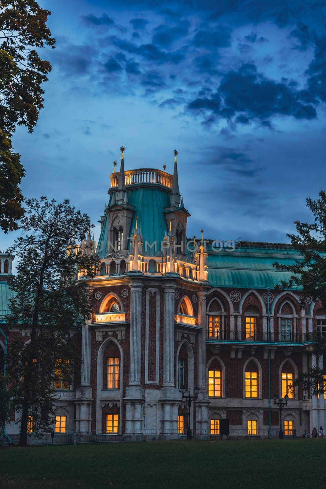A grand palace with Gothic architectural features, illuminated by warm lights, surrounded by trees under a twilight sky.