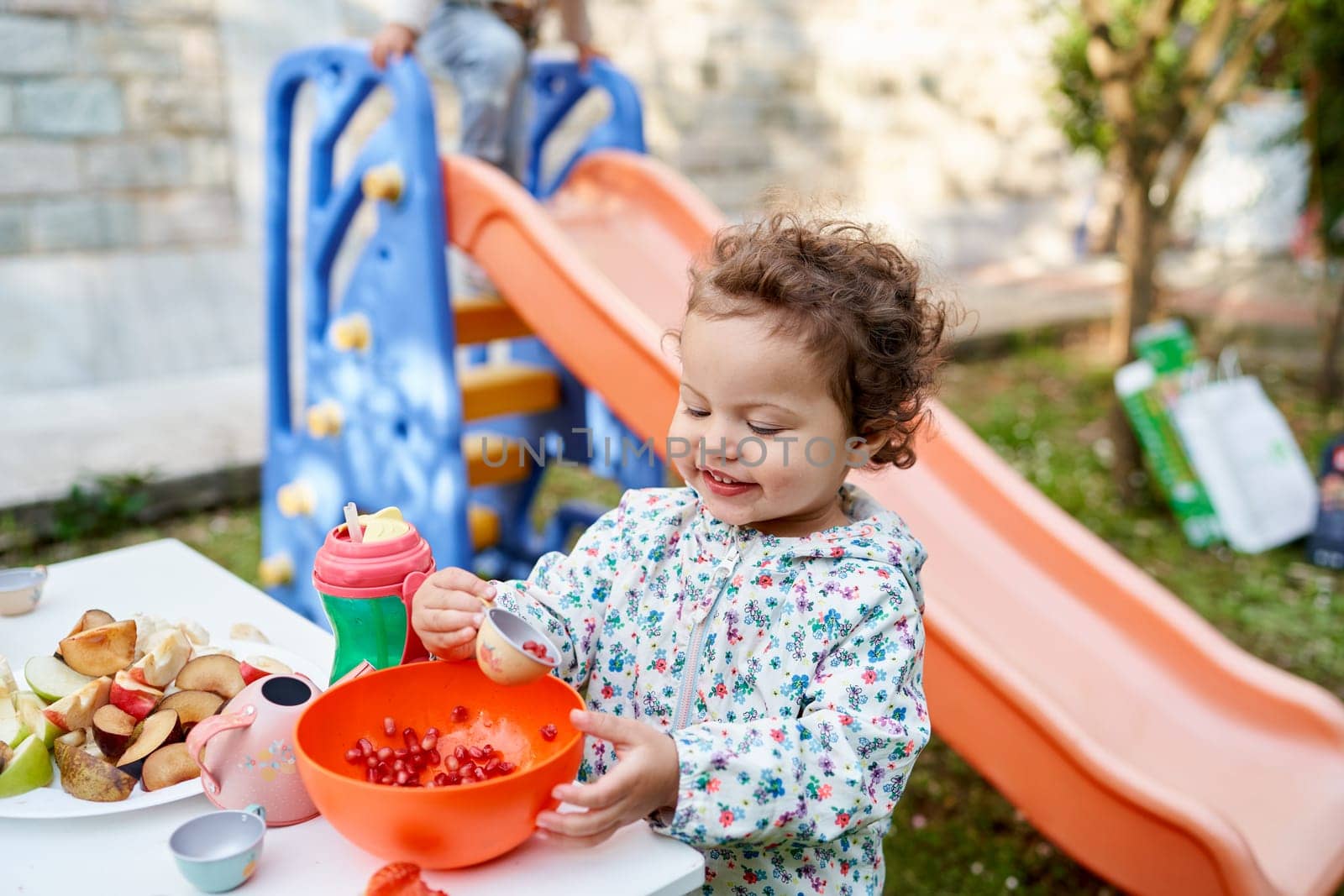 Little girl looks at pomegranate seeds in a small cup while standing at a festive table in the garden. High quality photo