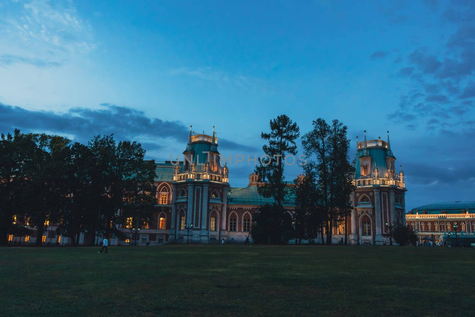 Wide view of a grand Gothic palace with illuminated towers, warm glowing windows, and surrounding trees under a twilight sky.
