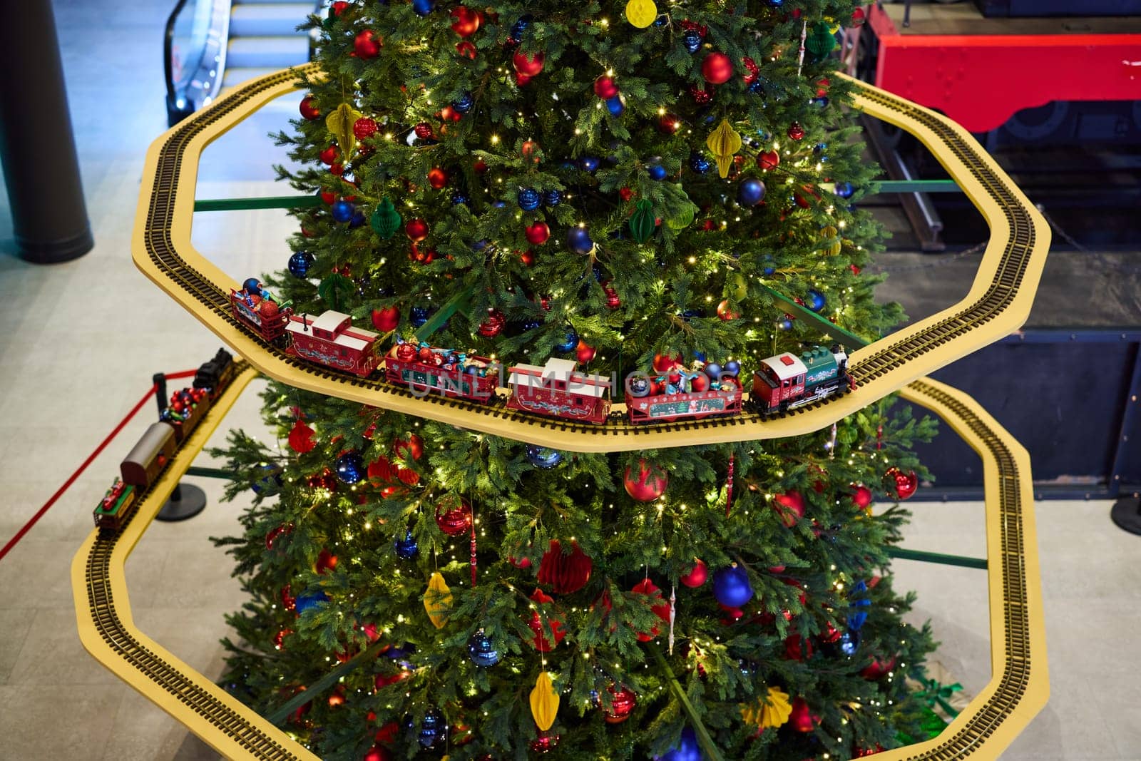 A colorful Christmas tree, adorned with ornaments, features a toy train set circling around its base