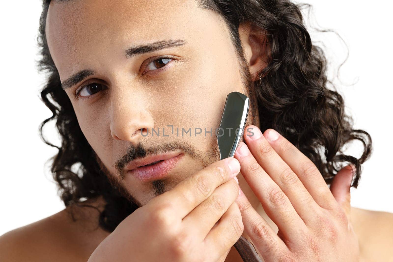 Man with curly hair grooming his beard carefully using a straight razor against a plain white background close up