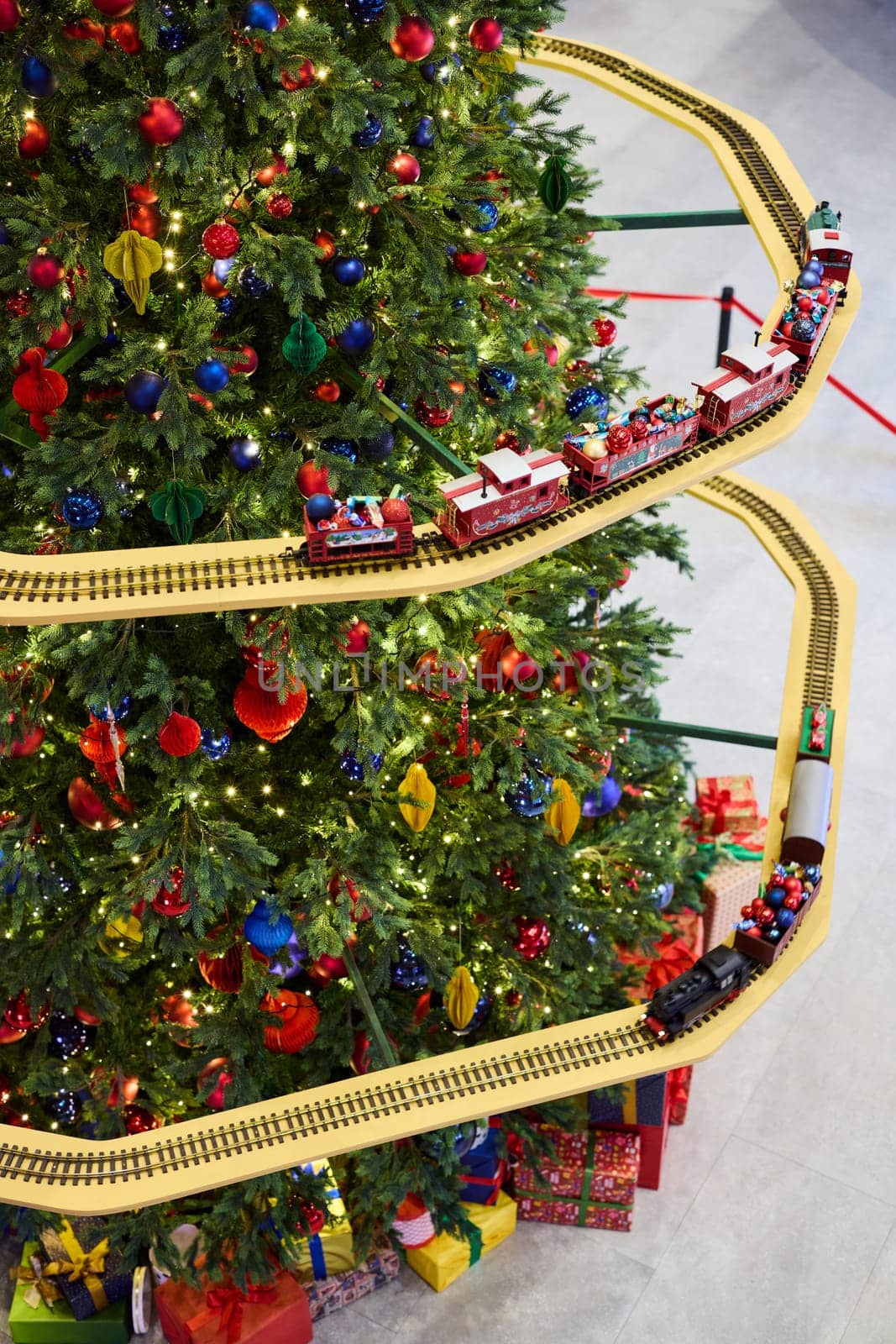 A beautifully vibrant Christmas tree is adorned with colorful, shiny ornaments and has a festive train track around it