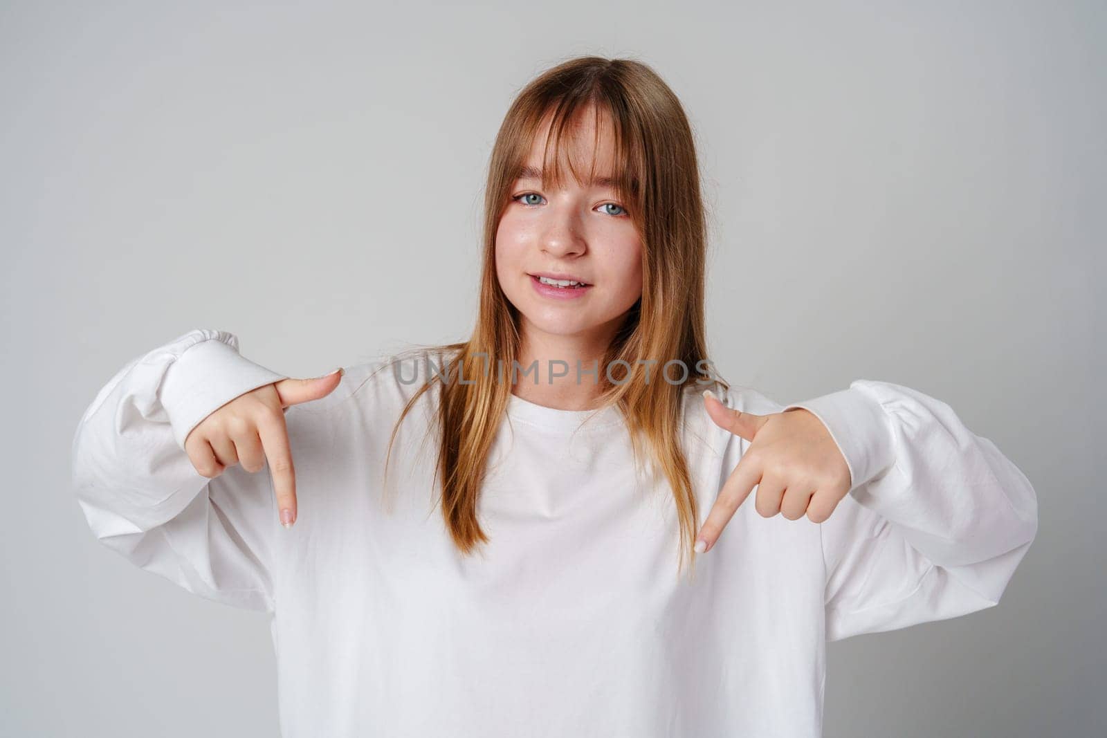 A young girl in a white long-sleeve shirt smiles while gesturing downwards in a light-colored room close up
