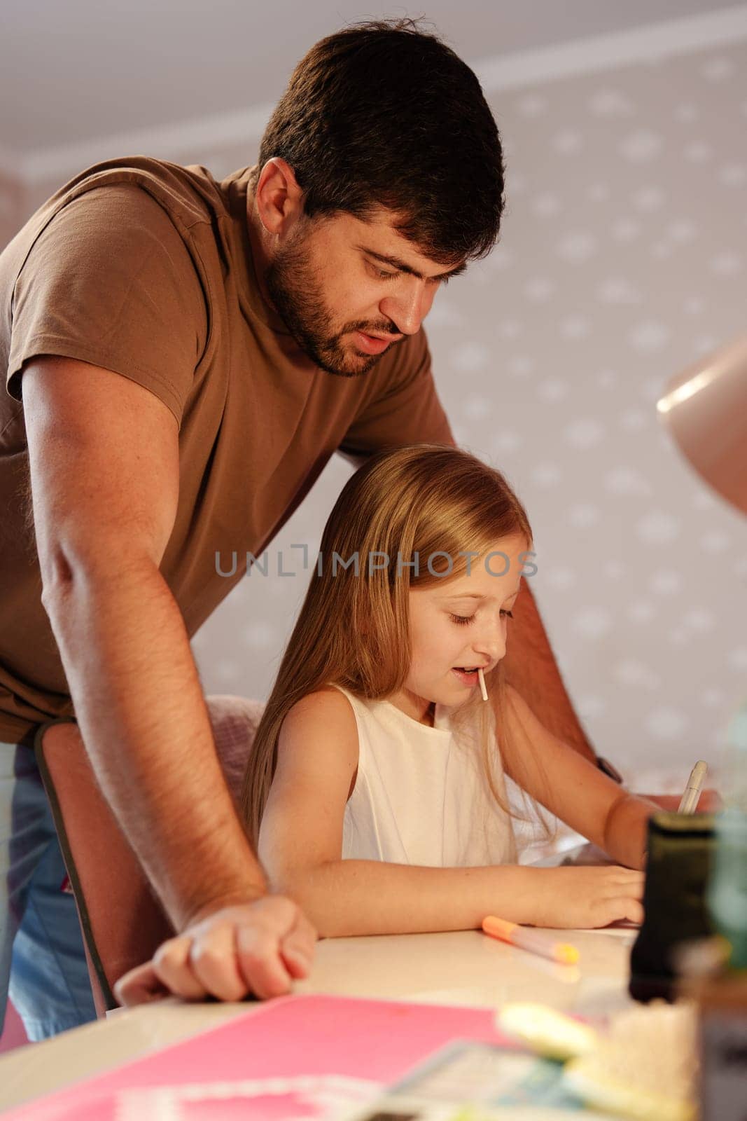 A father stands over his young daughter as she sits at a table working on her homework. He is looking down at her, watching her as she uses a pencil and paper.