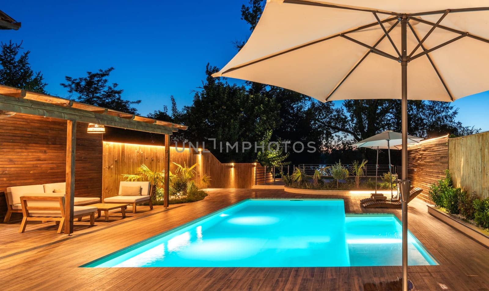 Backyard swimming pool with wooden deck and large umbrella in cozy ambiance. Luxury outdoor relaxation area with soft evening lighting