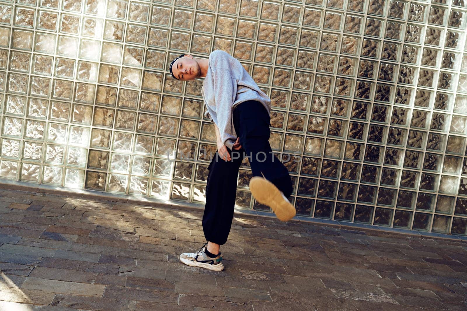 A young man in a gray sweatshirt and black pants performs a dance move in front of a wall made of glass blocks. His right foot is raised high in the air, demonstrating his flexibility and athleticism.