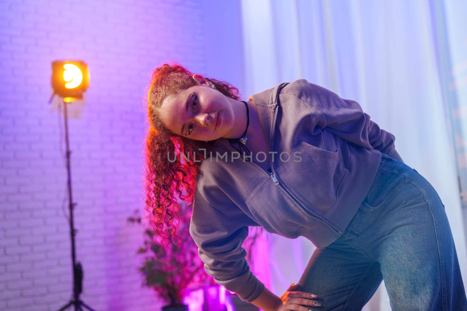 A young woman with long, pink curly hair poses in a studio, wearing a gray sweatshirt and blue jeans. She is bent at the waist, looking at the camera.