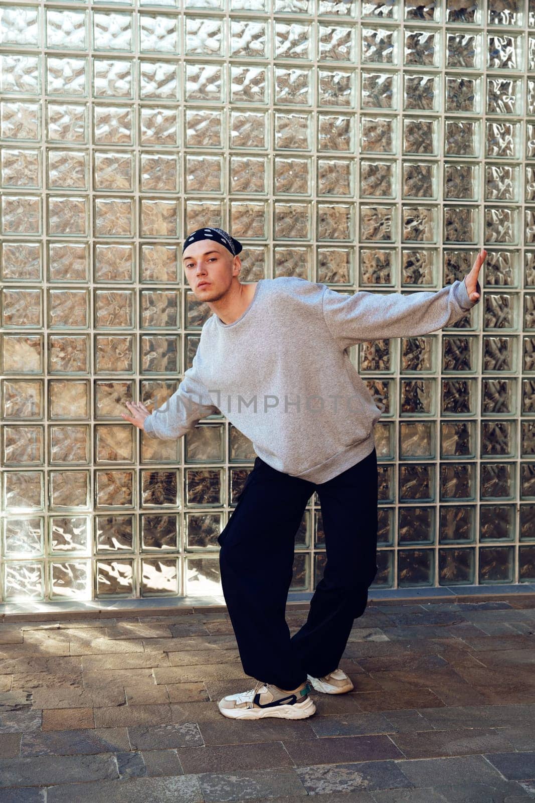 A man stands in front of a wall of glass blocks. He is wearing a grey sweatshirt, black pants, and white sneakers. He is looking at the camera and has his right arm outstretched.