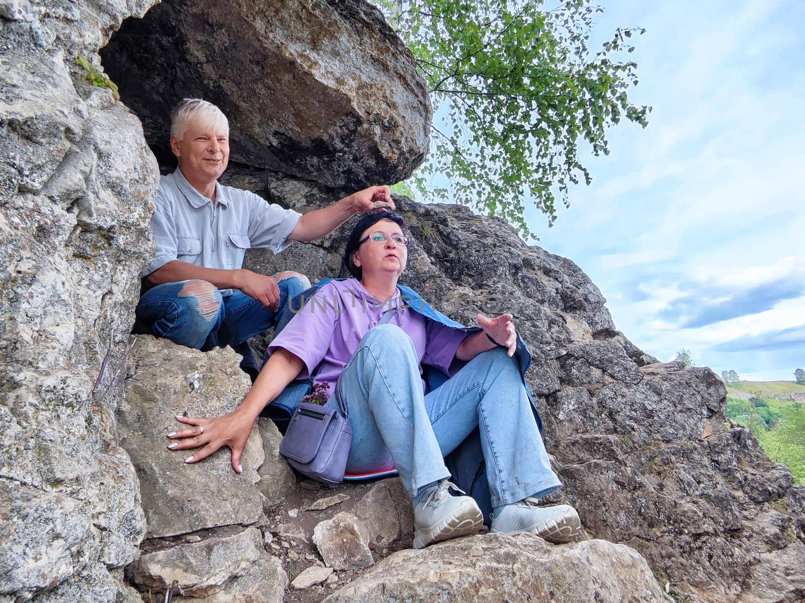 Two people relax in a rocky alcove, taking in the fresh air and surrounding greenery on a pleasant day.