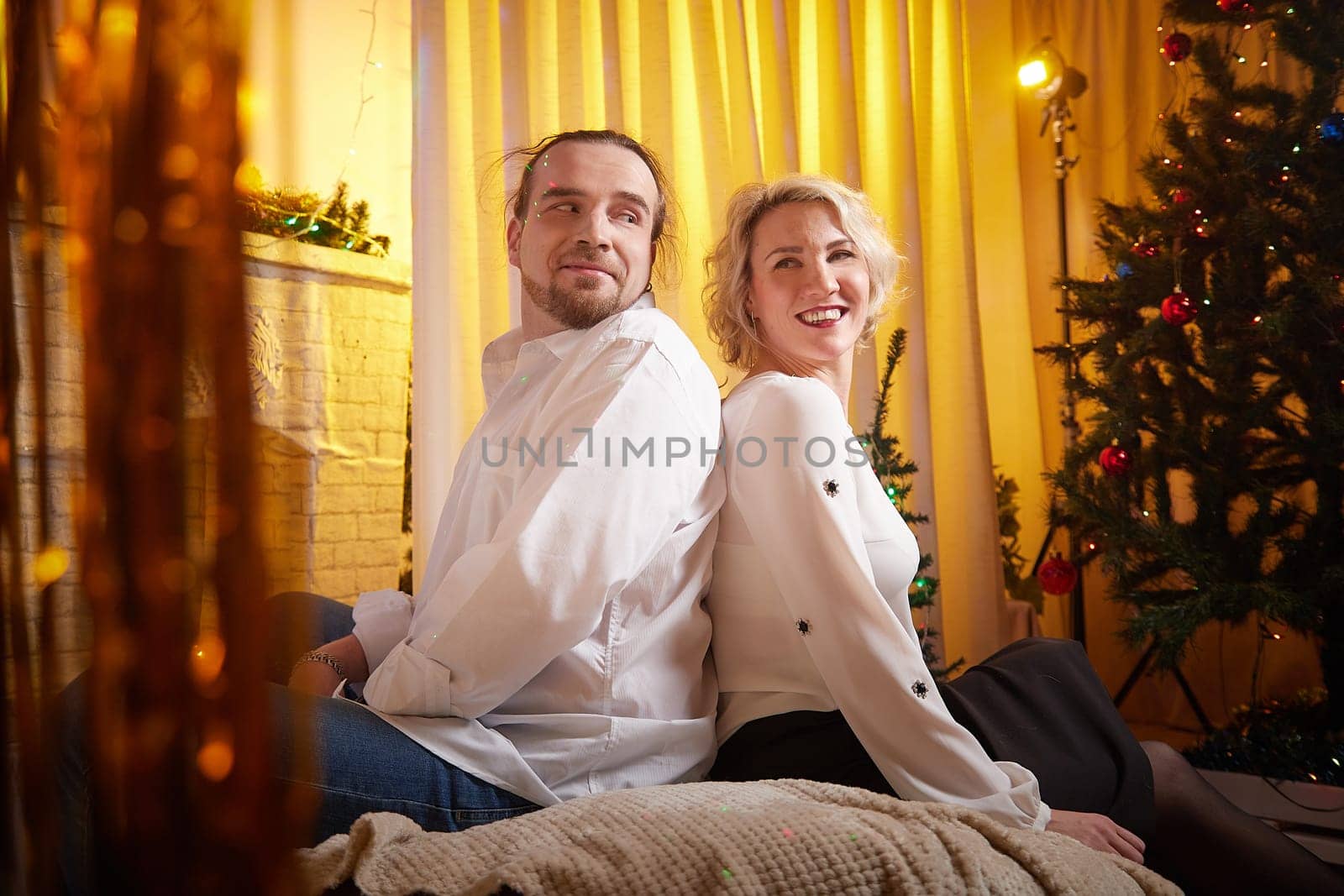 A couple sits back to back, smiling joyfully in a warmly lit room adorned with festive decorations and a Christmas tree.