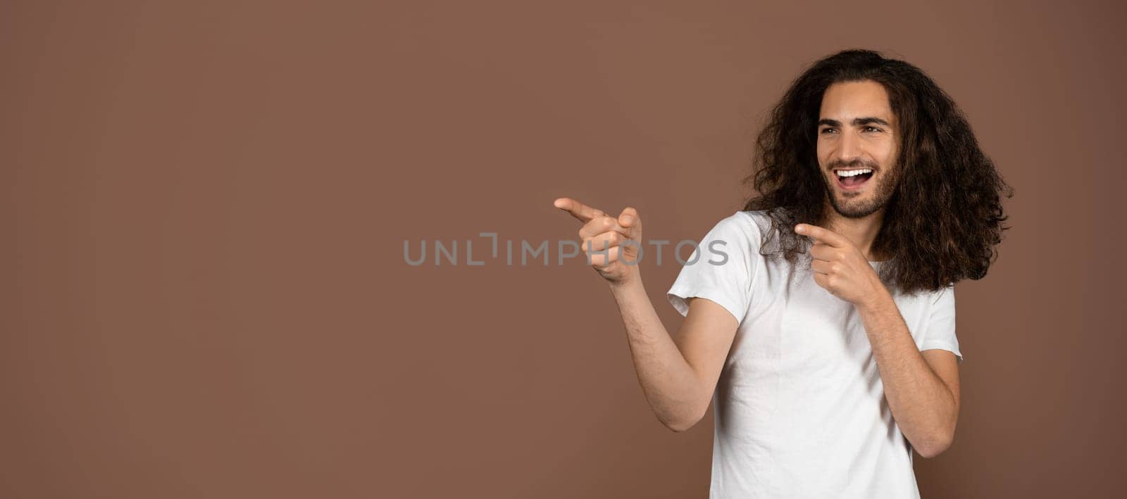 A young Caucasian man with long, curly hair joyfully poses while pointing at copy space. His carefree expression reflects a vibrant and playful attitude toward life.