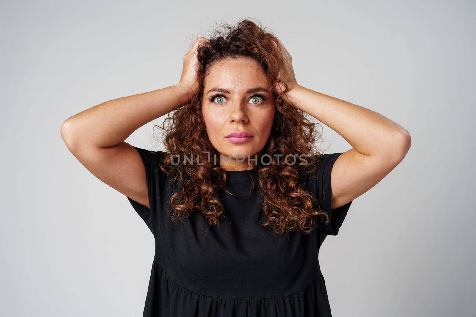 Smiling woman with curly hair poses confidently against a plain background while holding her hands behind her head indoors