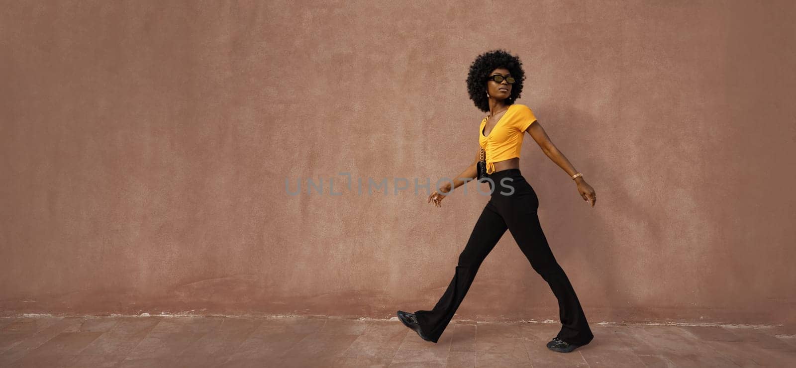 A trendsetter strolls effortlessly along a brown wall, showcasing a bright yellow top paired with black flared pants. The confident posture and cool sunglasses enhance their fashion statement.