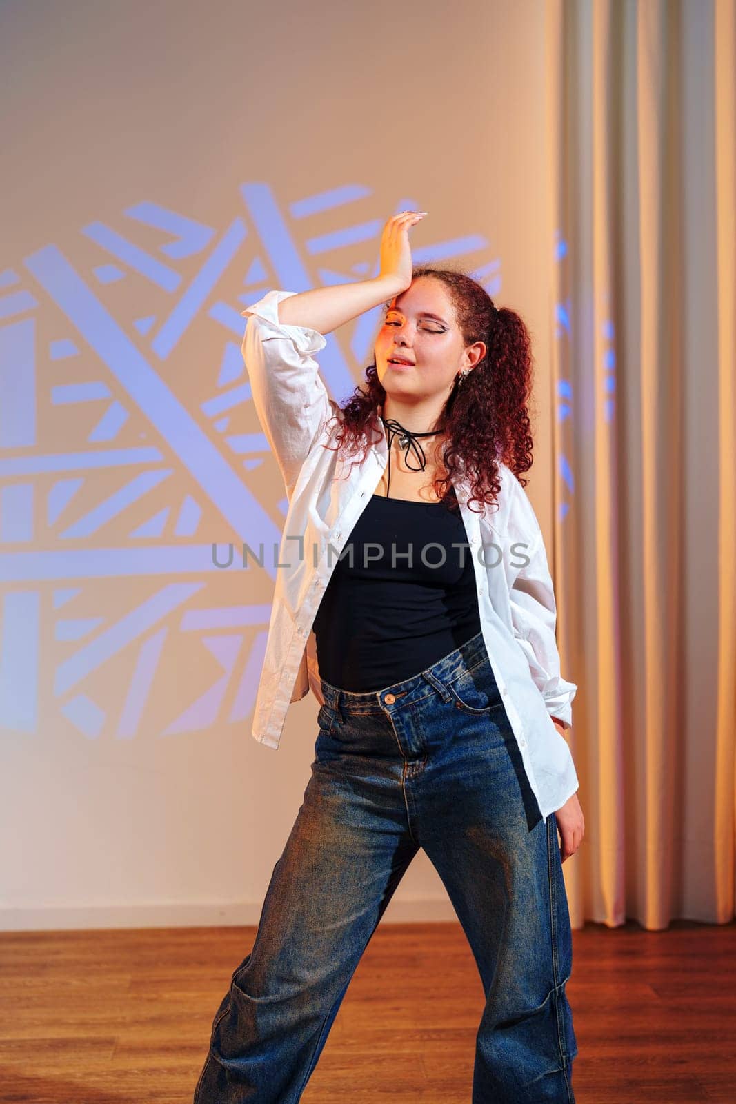 A young woman with long, curly red hair is standing in a dance studio. She is wearing a white button-up shirt over a black tank top and blue jeans.