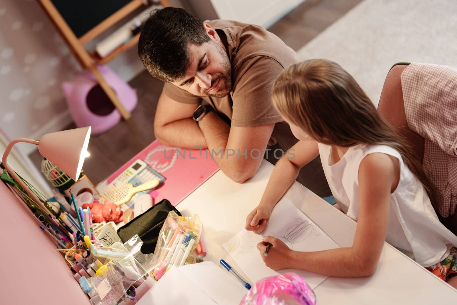 A father and daughter are sitting at a desk together. The daughter is drawing on a piece of paper while the father looks on. There are many school supplies on the desk.