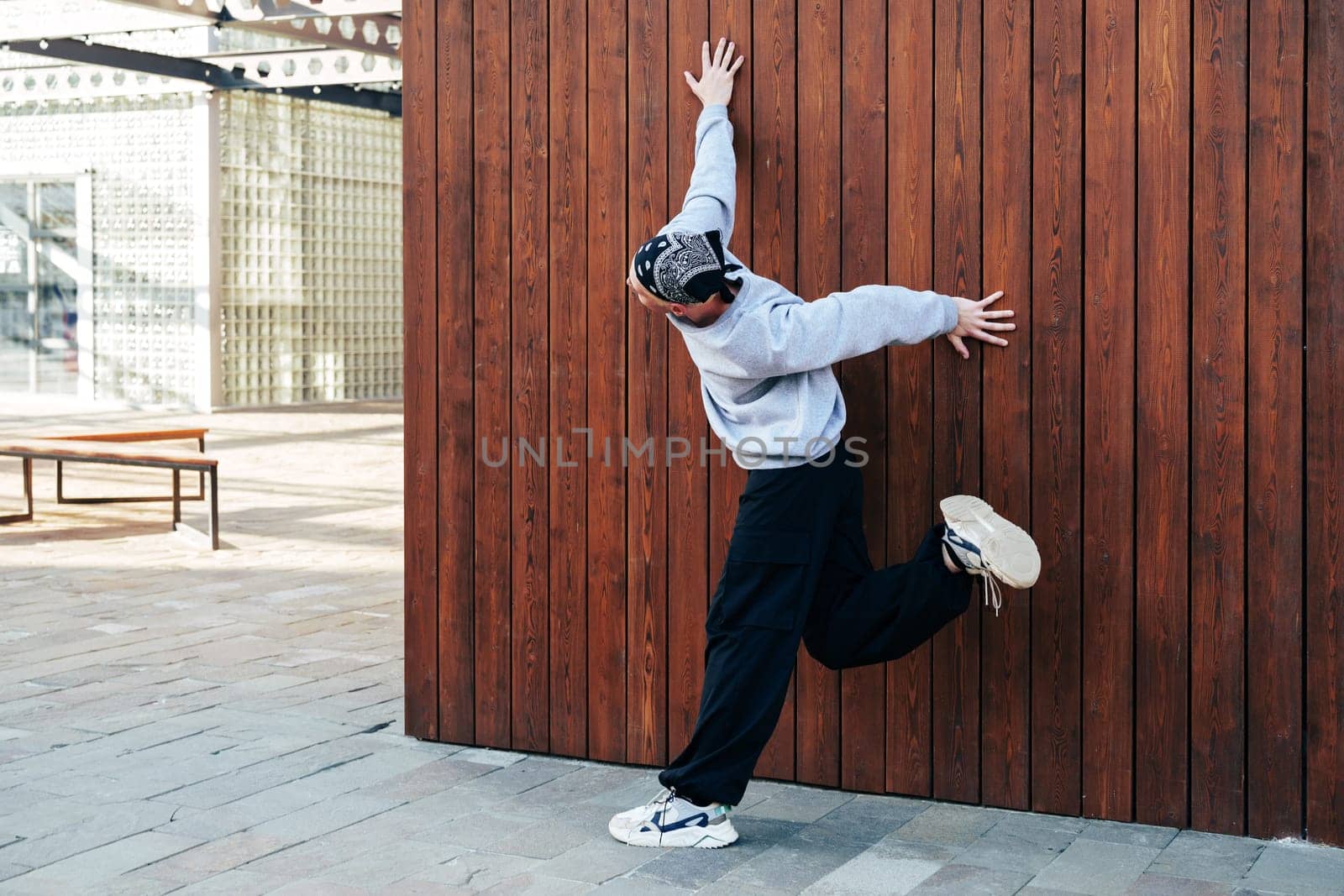 A man in a grey sweatshirt and black pants stands on a pavement, facing a wooden wall, with one hand touching the wall and his leg raised behind him.