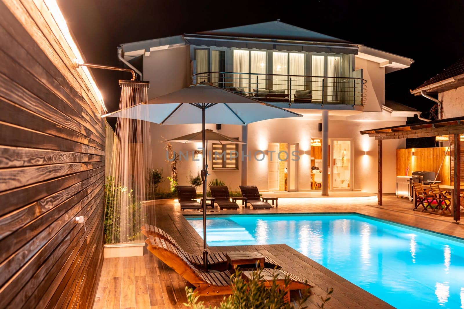 Luxury villa with illuminated pool, wooden deck, and outdoor shower at night. Concept of modern backyard design