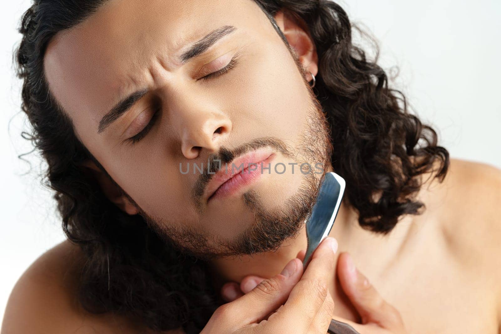 Man with curly hair grooming his beard carefully using a straight razor against a plain white background close up