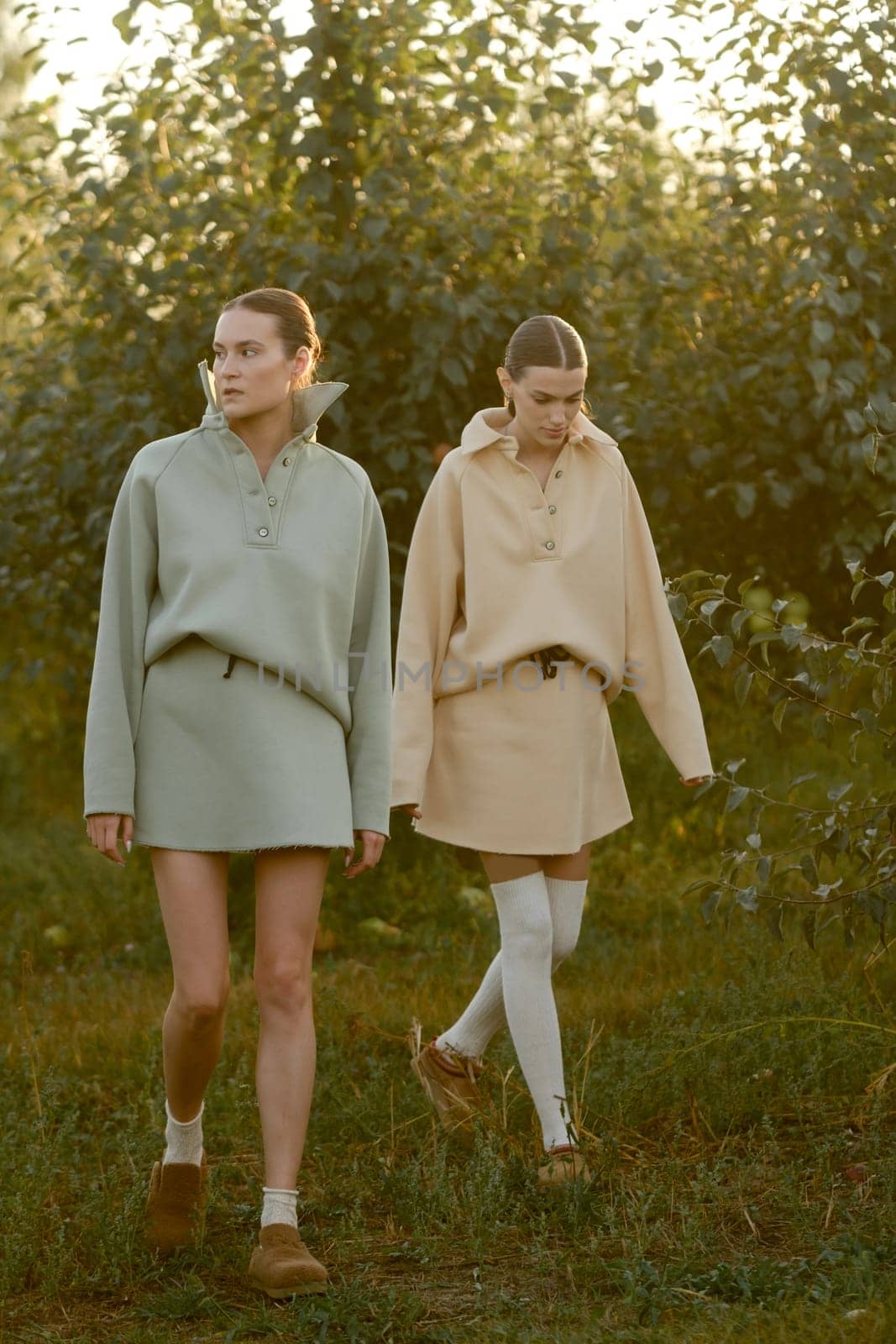 Two models in stylish tracksuits pose in apple orchard at sunset . The model is wearing a light-colored tracksuit and skirt.