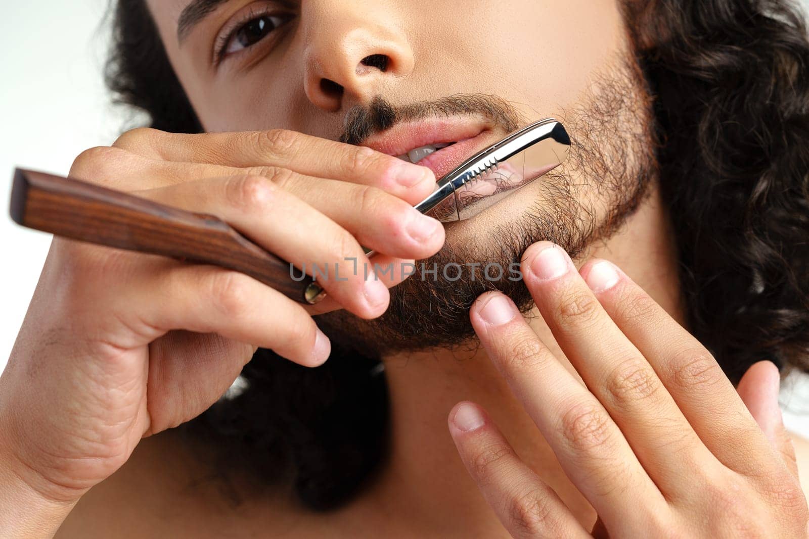 Man with curly hair grooming his beard carefully using a straight razor against a plain white background close up