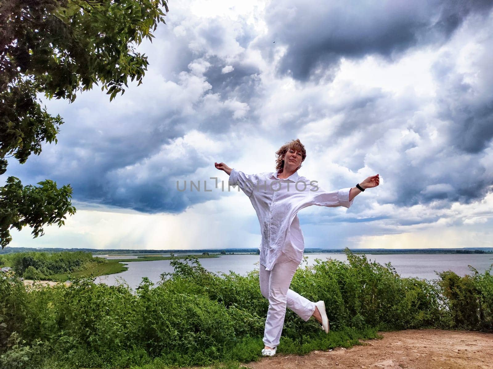 Blonde woman in white outfit celebrates at a river viewpoint, enjoying nature and the moment with an adventurous spirit.