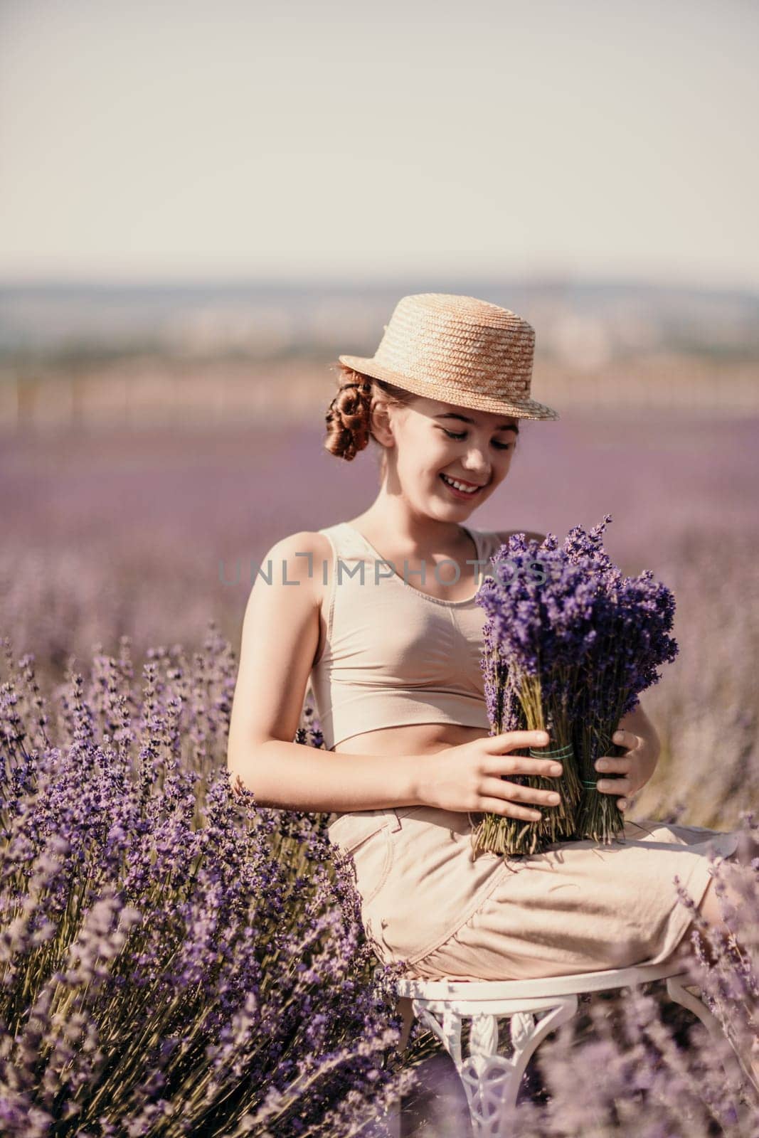 A woman is sitting in a field of lavender flowers and wearing a straw hat. She is smiling and holding a bouquet of flowers. Scene is peaceful and serene, as the woman is surrounded by the beauty of nature.