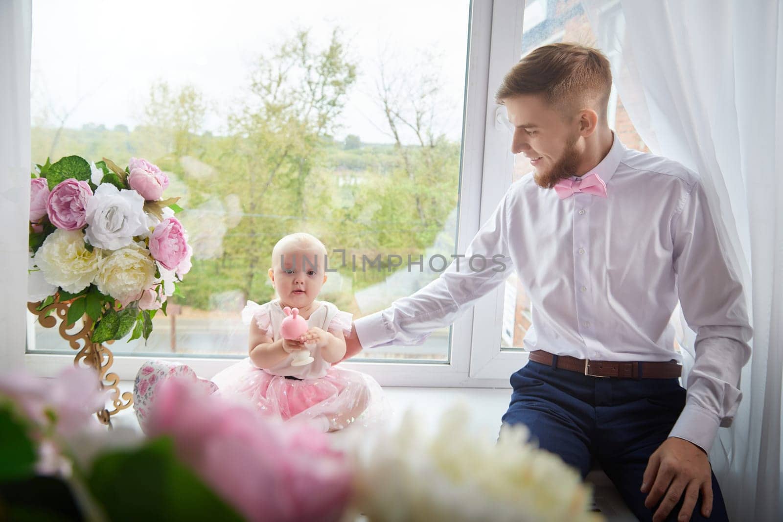 A man is carefully holding a baby who is dressed in a bright pink dress in the window in room with flowers. Photoshoot with happiness moment
