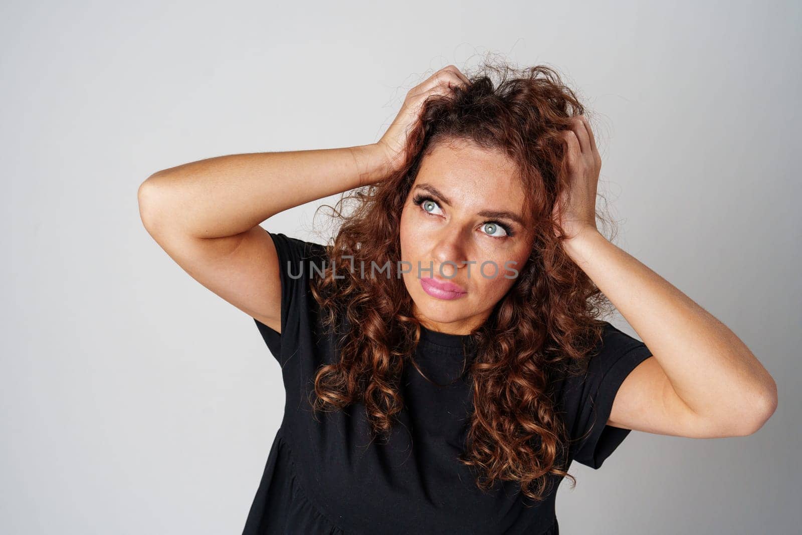 Smiling woman with curly hair poses confidently against a plain background while holding her hands behind her head indoors