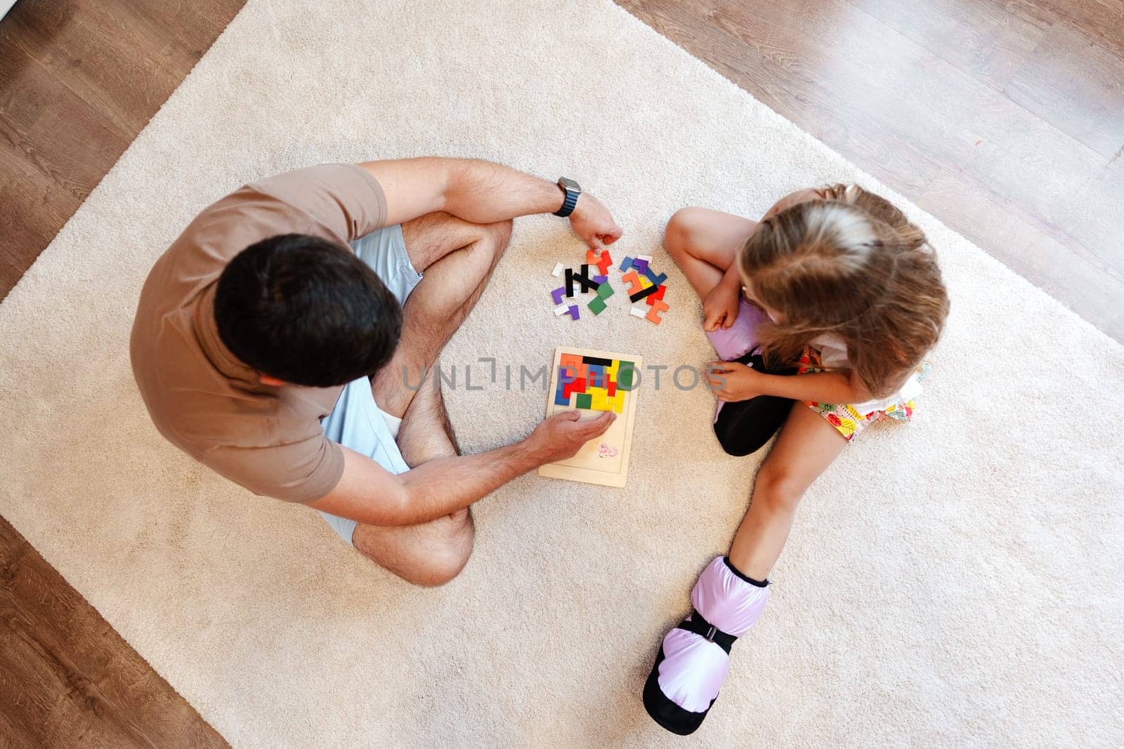 A man and a young girl sit on a white rug and work together on a colorful puzzle. The man is wearing a tan t-shirt and shorts, and the girl is wearing a yellow and white dress.