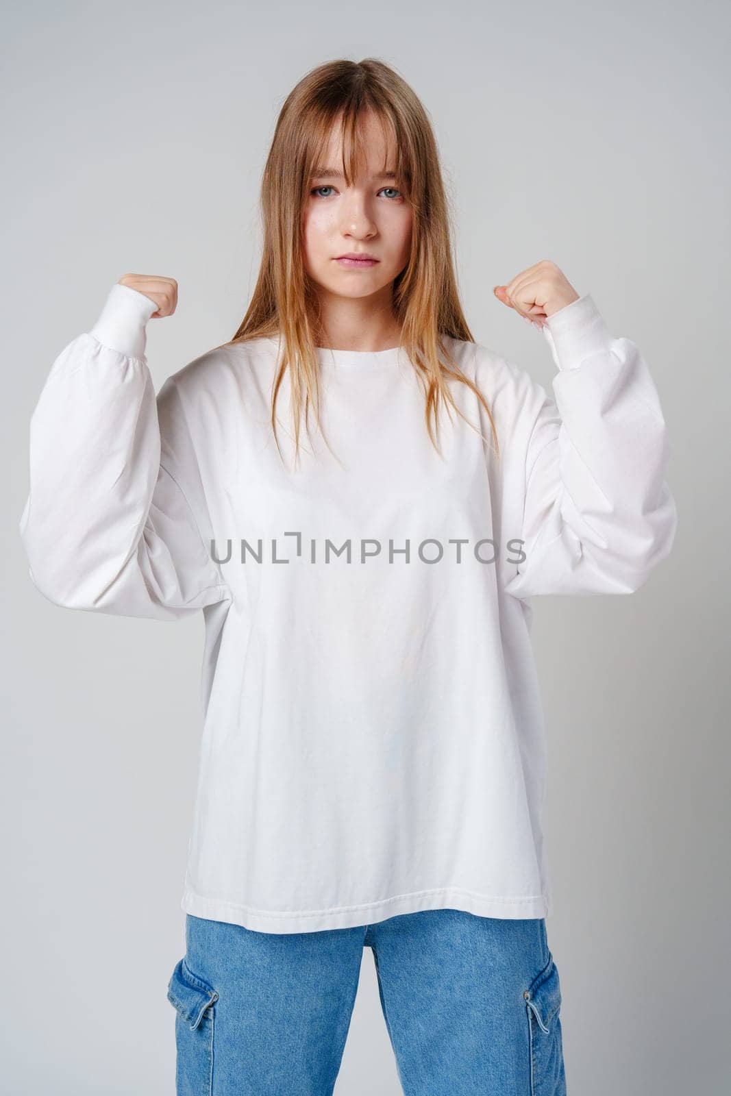 A joyful young girl with long hair wearing a white sweater stands against a plain background close up