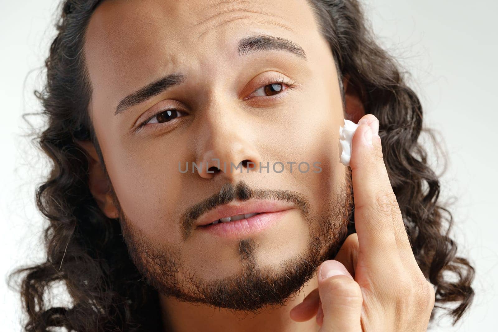 A young man applying skincare cream on his face with a focused expression, demonstrating personal grooming at home close up