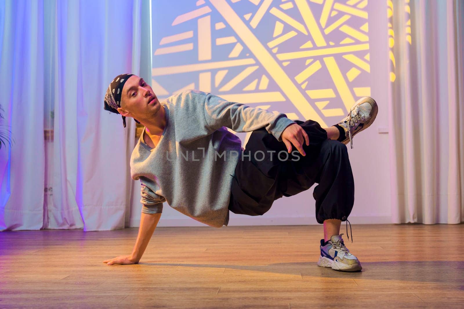 A dancer in a gray sweatshirt and black pants performs a breakdancing move on a wooden floor. Behind the dancer are white curtains and a light projection with a geometric pattern on the wall.
