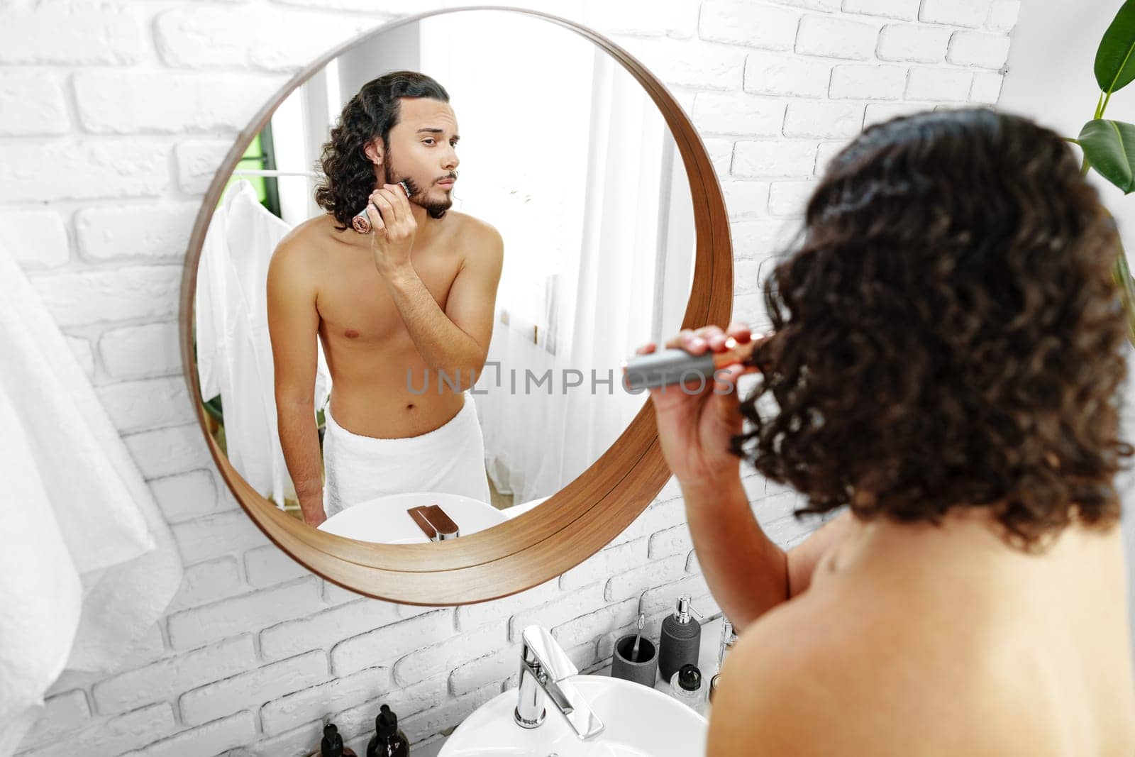 A man with curly hair grooming himself in the bathroom while looking into a round mirror close up